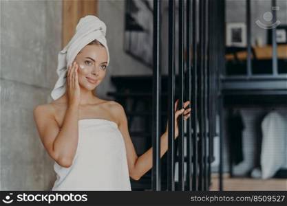 Beautiful healthy young woman applies face cream on complexion, looks thoughtfully somewhere into distance, wrapped in towels, stands indoor, has perfect pure skin. Women and cosmetology concept
