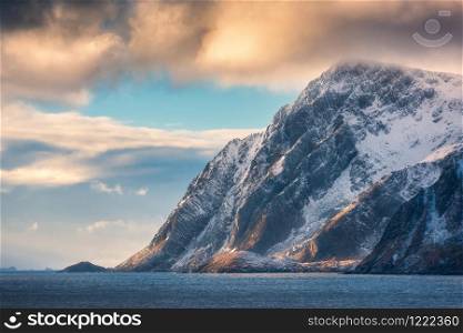 Beautiful high snow covered mountains at sunset in winter. Lofoten Islands, Norway. Landscape with snowy rocks, sea, blue sky with orange clouds in the evening. Travel and nature