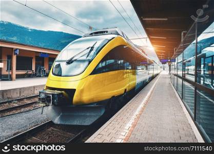 Beautiful high speed train on the railway station in mountains at sunset in autumn Yellow modern commuter train on the railway platform. Industrial landscape with railroad. Passenger transportation
