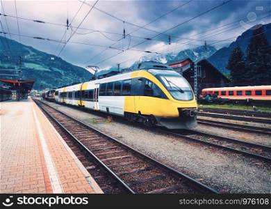 Beautiful high speed train on the railway station in mountains at sunset in autumn Yellow modern commuter train on the railway platform. Industrial landscape with railroad. Passenger transportation
