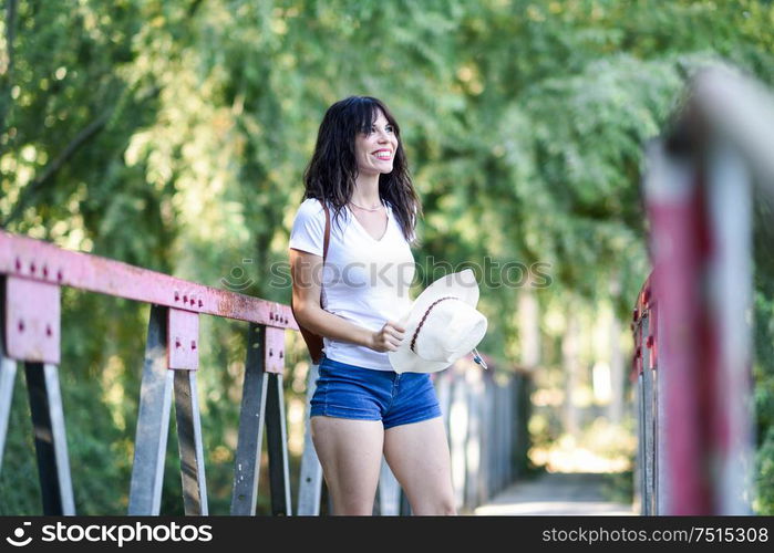 Beautiful hiker young woman with backpack, with blue eyes, wearing straw hat, hiking in the countryside and having fun on a rural bridge.. Woman with backpack standing on rural bridge.