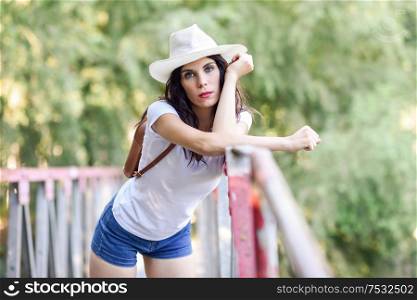 Beautiful hiker young woman with backpack, with blue eyes, wearing straw hat, hiking in the countryside and having fun on a rural bridge.. Woman with backpack standing on rural bridge.
