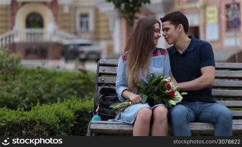 Beautiful hipster couple in love sitting on bench in park during romantic date. Smiling cute girl with bunch of flowers sitting on bench while handsome man whispering romantic words, expressing feelings and affection, sharing good emotion.