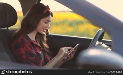 Beautiful hipster female driver sitting at steering wheel and surfing the net with mobile phone while travelling by car on summer vacation. Charming woman using smartphone while sitting in driver&acute;s seat during road trip on countryside. Side view.