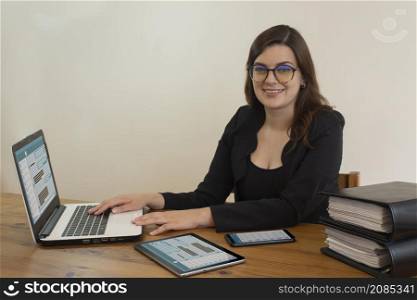 Beautiful Hispanic female office worker with glasses smiling happy sitting at her desk working in front of her computer against white wall. Beautiful Hispanic female office worker with glasses happy sitting at her desk working in front of her computer