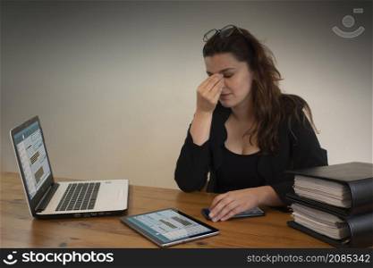 Beautiful Hispanic female office worker with headache massaging her eyes with her left hand, sitting at her desk in front of her computer. Beautiful Hispanic female office worker massaging her eyes with her left hand, sitting at her desk in front of her computer