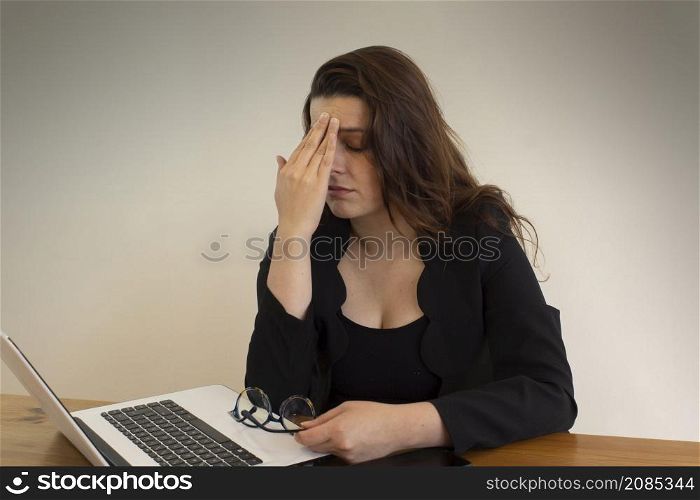 Beautiful Hispanic female office worker with headache massaging her eyes with her left hand, sitting at her desk in front of her computer. Beautiful Hispanic female office worker massaging her eyes with her left hand, sitting at her desk in front of her computer