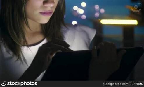 Beautiful hispanic girl typing on touch screen device, sitting at home at night. The young woman is studying on her tablet and is concentrated