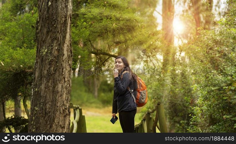 Beautiful Hispanic woman dressed in black with backpack walking alone on a forest path with sun rays between the trees during the morning. Beautiful Hispanic woman dressed in black with backpack walking alone on a forest path with sun rays between the trees