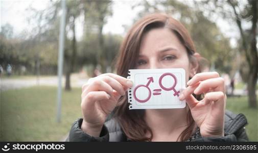 Beautiful Hispanic woman in the middle of a park holding in her hand a paper with the message of equality with the icons of man and woman in the foreground. Beautiful Hispanic woman in the middle of a park holding in her hand a paper with the message of equality with the icons of man and woman