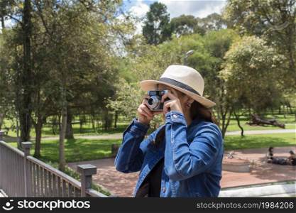 Beautiful Hispanic woman with a hat taking a picture with an old camera in the middle of a park on a sunny morning. Beautiful Hispanic woman taking a picture with an old camera in the middle of a park on a sunny morning