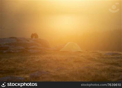 Beautiful image of unidentified young couple wild camping in English countryside during stunning Summer sunrise with warm glow of the sun lighting the landscape
