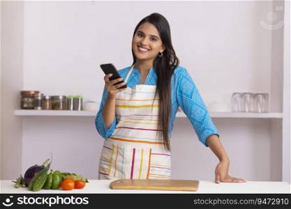 Beautiful Indian woman looking at camera with holding smartphone in Kitchen 