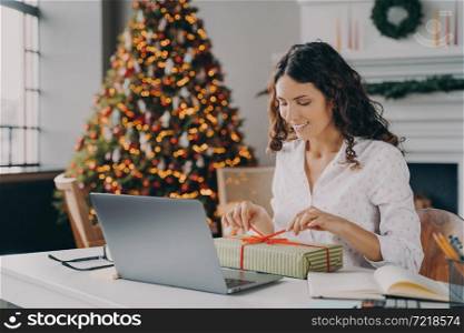 Beautiful Italian woman cheerful business lady decorating Xmas present with colored ribbon while sitting at desk at modern home office against backdrop of festive Christmas tree full of bright lights. Beautiful Italian woman business lady decorating Xmas present at workplace during Christms holidays