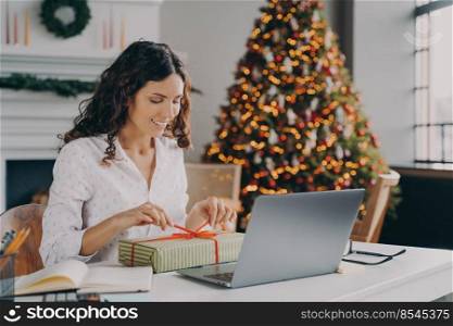 Beautiful Italian woman cheerful business lady decorating Xmas present with colored ribbon while sitting at desk at modern home office against backdrop of festive Christmas tree full of bright lights. Beautiful Italian woman business lady decorating Xmas present at workplace during Christms holidays