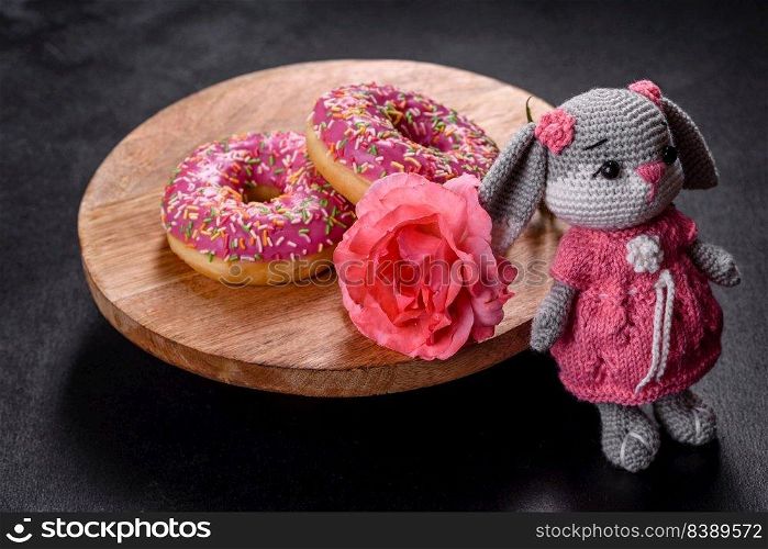 Beautiful knitted rabbit toy with doughnuts with pink glaze and coloured sprinkle on a dark concrete background. Beautiful knitted rabbit toy with doughnuts with pink glaze and coloured sprinkle