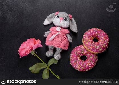 Beautiful knitted rabbit toy with doughnuts with pink glaze and coloured sprinkle on a dark concrete background. Beautiful knitted rabbit toy with doughnuts with pink glaze and coloured sprinkle