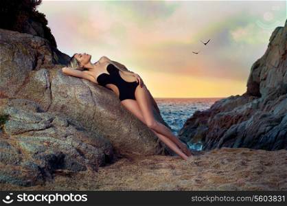 Beautiful lady in black swimsuit on rocky shore. Sunset travel photo
