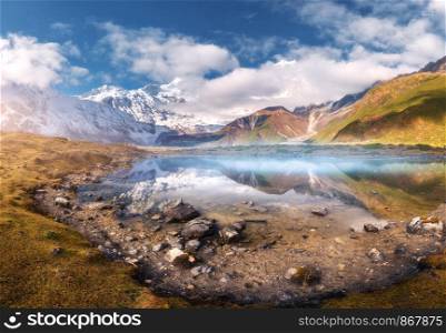 Beautiful lake with fog in mountain valley at sunrise in autumn. Landscape with snowy Manaslu mountain, stones, yellow grass, green hills, blue sky with clouds, reflection in water in Nepal. Nature