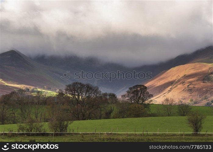Beautiful landscape image of Autumn in Lake District looking across to mountains in distance with sunlight dappled across fields