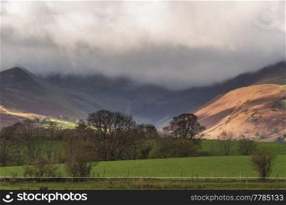Beautiful landscape image of Autumn in Lake District looking across to mountains in distance with sunlight dappled across fields