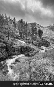 Beautiful landscape image of Ogwen Valley river and waterfalls during Winter with snowcapped mountains in background in black and white