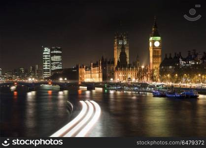 Beautiful landscape image of the London skyline at night looking. England, London, London. View along River Thames at night towards Houses of Parliament and Big Ben with light streaks from a boat.. Landscape image of the London skyline at night looking along the River Thames