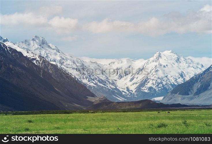 Beautiful landscape of mountain ranges and green grass meadow field under white clouds in the blue sky in sunny summer day. Shot in Mt Cook, the highest mountain in New Zealand.