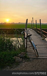 Beautiful landscape of nature with lovely orange sunset, bicycle put up on wooden fence, small wooden bridge across a river and in the distance is green rice field