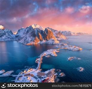 Beautiful landscape with blue sea, snowy mountains, rocks and islands, village, rorbu, road, bridge and pink sky. Aerial view. Hamnoy in snow at sunrise in winter in Lofoten islands, Norway. Top view