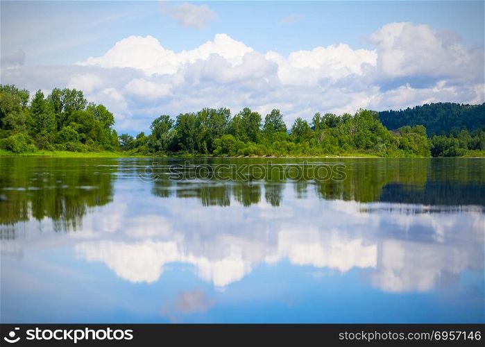 Beautiful landscape with blue sky and white clouds reflected in the clear river water. Wooded waterside of a mountain lake. Summer idyllic landscape.. Beautiful landscape with blue sky and white clouds reflected in