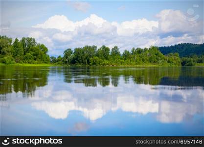 Beautiful landscape with blue sky and white clouds reflected in the clear river water. Wooded waterside of a mountain lake. Summer idyllic landscape.. Beautiful landscape with blue sky and white clouds reflected in
