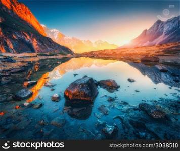 Beautiful landscape with high mountains with illuminated peaks, stones in mountain lake, reflection, blue sky and yellow sunlight in sunrise. Nepal. Amazing scene with Himalayan mountains. Himalayas. Mountains with illuminated peaks, stones in mountain lake at sun