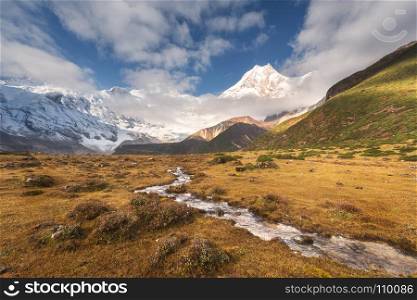 Beautiful landscape with high mountains with snow covered peaks, small river, yellow grass and cloudy sky at colorful sunrise. Mountain valley. Nepal. Amazing scene with Himalayan mountains. Nature. Mountains with snow covered peaks and small river
