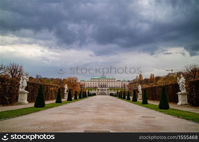 Beautiful landscape with wide main walking alley before Schloss Belvedere Palace with regular planting of trees and ancient statues in Vienna, Austria on a background of cloudy sky.. View of Schloss Belvedere Palace with walking alley and ancient statues in Vienna.