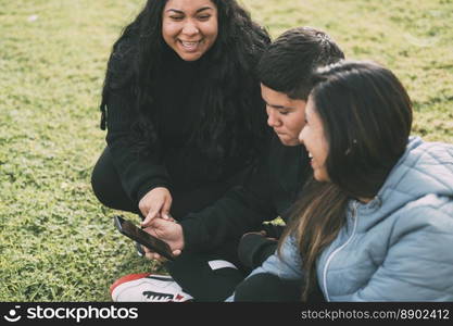 beautiful latin family laughing from watching smartphone videos, sitting on the green park two brothers and the mother on a sunny day.. group of Latinos laughing sitting on the ground in a park with a smartphone