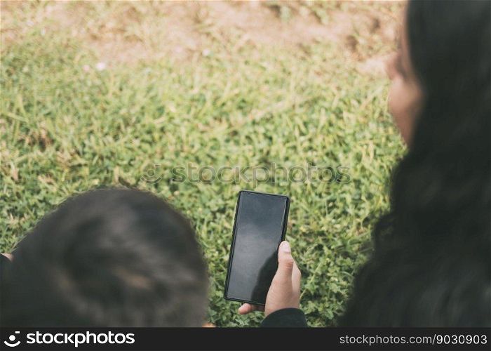 beautiful latin family laughing from watching smartphone videos, sitting on the green park two brothers and the mother on a sunny day.. view from the back group of laughing Latinos sitting on the ground in a park with a smartphone