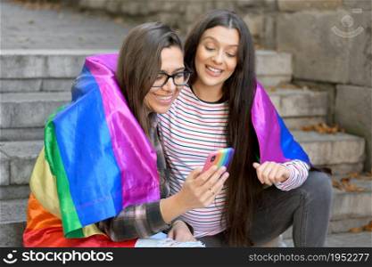 Beautiful lesbian couple with rainbow flag using a mobile phone in the street. High quality photo. Beautiful lesbian couple with rainbow flag using a mobile phone in the street.