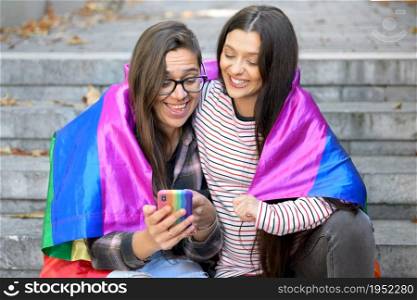 Beautiful lesbian couple with rainbow flag using a mobile phone in the street. High quality photo. Beautiful lesbian couple with rainbow flag using a mobile phone in the street.
