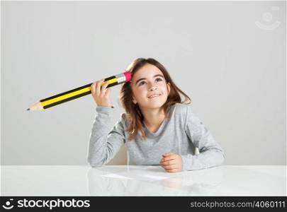Beautiful little girl in a desk playing with a big pencil, against a gray background