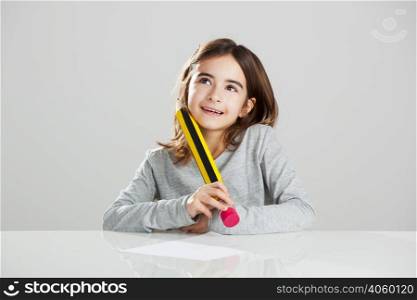 Beautiful little girl in a desk playing with a big pencil, against a gray background