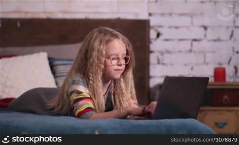 Beautiful little girl playing with a laptop on the bed. Gracious little blonde princess in glasses with long hair and blue eyes playing the game on a laptop computer and smiling joyfully in modern room.