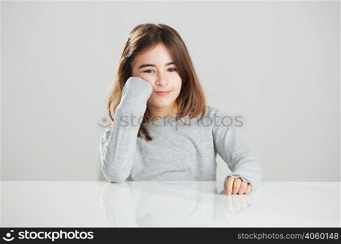 Beautiful little girl sitting behind a desk and smiling, against a gray background