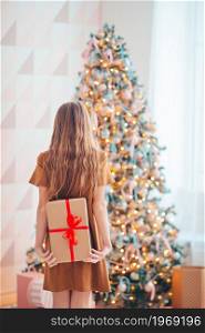 Beautiful little girl with a gift. Back view of kid holds a gift box near the Christmas tree indoors. Merry Christmas and Happy Holidays.. Back view of kid holds a gift box near the Christmas tree indoors.
