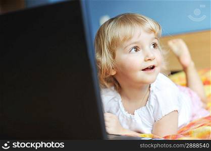 Beautiful little girl with laptop lying on bed at house