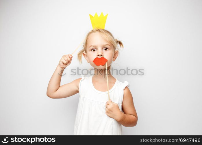 Beautiful little girl with paper crown and red lips posing on white background at home.. Beautiful little girl with paper crown and red lips posing on white background at home