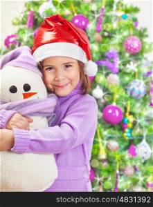 Beautiful little happy girl wearing red Santa hat and holding in hands snowman soft toy, Christmas tree, happy winter holidays