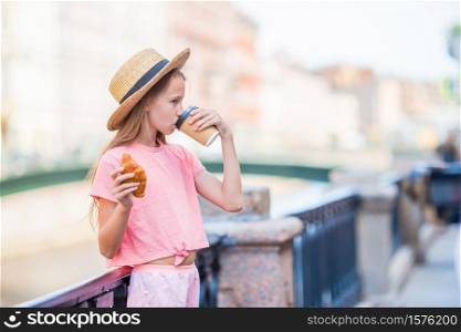 Beautiful little tourist girl with a croissant and coffee outdoors on the promenade on the river. Woman with a croissant and coffee outdoors on the promenade