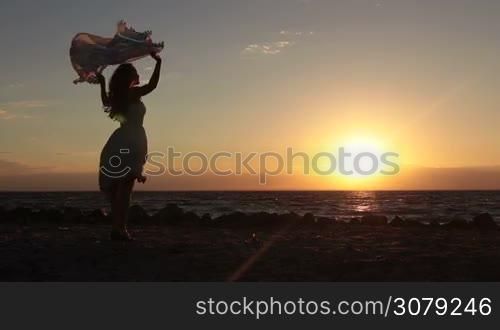 Beautiful long blonde hair female in summer dress with hands up holding silk scarf in wind on the beach in twilight time. Gorgeous young woman with flying scarf fluttering in the wind standing on seaside in glow of amazing yellow sunset.