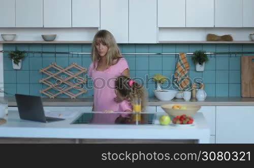 Beautiful long blonde hair woman working with laptop at home browsing bills and documents while little daughter drinking orange juice in kitchen. Busy mother working with laptop computer using online mortgage calculator to save money on home loan.